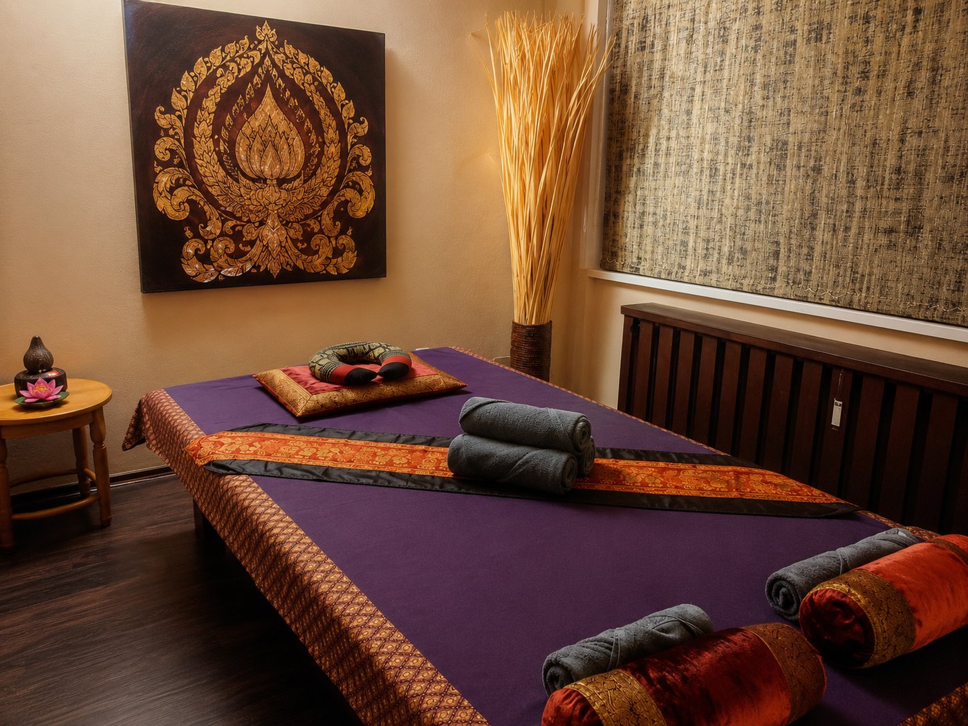 Behandlungsraum bei Thailin Thaimassage: Massageliege mit lila Auflage und Thai-Seidenläufern, gerollte Handtücher, im Hintergrund ein goldenes Lotusbild an der Wand.